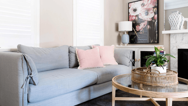 Bright, refined living room featuring a large pale blue upholstered sofa with pink cushions, a round wood-and-glass coffee table, a fireplace with a light stone surround, framed floral artwork, a table lamp, and indoor greenery. White plantation shutters and light neutral finishes create an airy, softly layered interior.