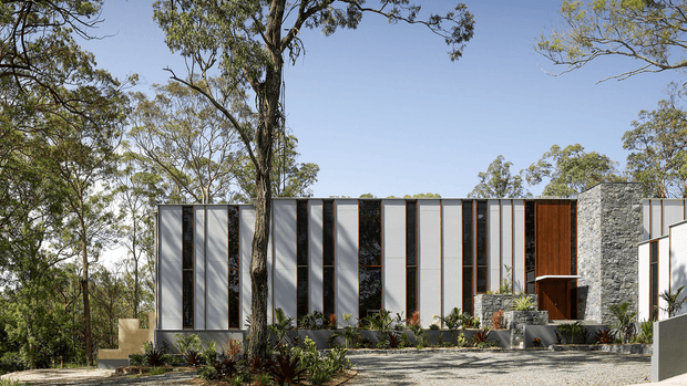 A contemporary low-rise residence set within a wooded landscape. The facade features a rhythmic composition of tall narrow glazing and light vertical wall panels, dark metal framing, warm timber-clad entry doors, and a prominent rough stone-clad volume. A gravel forecourt, planted garden beds, and mature eucalyptus trees complete the exterior setting.