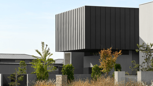 Exterior architectural photograph of a contemporary two-storey residence featuring a bold cantilevered upper volume clad in dark vertically ribbed panels, contrasting with light rendered walls and a pale metal roof section. The property is enclosed by a dark slatted fence with low masonry and rendered garden walls, landscaped with ornamental grasses and small trees.