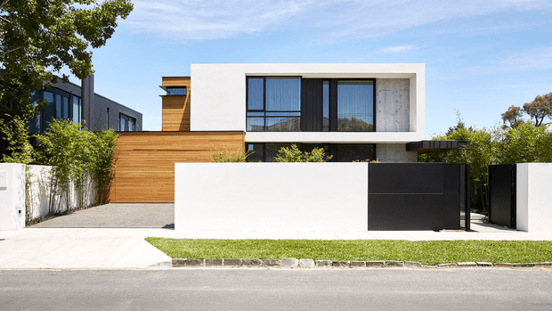 Street-facing view of a contemporary multi-level residence with a crisp white rectangular upper volume, extensive dark-framed glazing, warm horizontal timber cladding at ground level, and a contrasting black garage or entry gate. A low white and black boundary wall, paved driveway, and neatly maintained lawn complete the modern suburban facade.