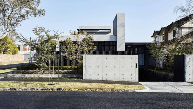 Street-facing view of a contemporary multi-level residence with a strongly geometric composition. The facade combines smooth pale concrete walls, dark vertical screening, black-framed glazing, a flat roofline, and a tall rectangular concrete chimney or feature volume. A gated driveway and minimalist landscaped front garden sit behind a low concrete boundary wall, with neighbouring homes visible at the edges.