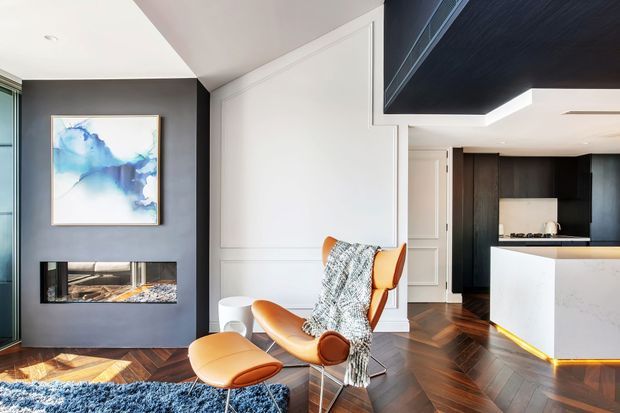 Contemporary open-plan living space with a sculptural tan leather lounge chair and ottoman, dark charcoal fireplace wall, framed artwork, white decorative wall panelling, and a compact kitchen with dark cabinetry and a white island. The room features richly patterned dark timber herringbone flooring, layered recessed and cove lighting, and a refined neutral palette with warm wood and leather accents.