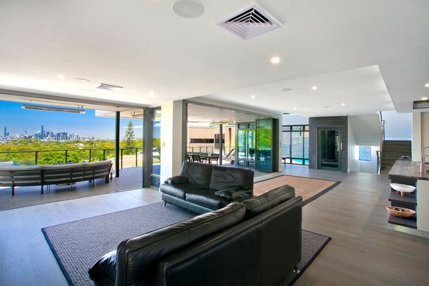 Bright contemporary open-plan living room with black leather sofas, a large textured rug, and grey-toned flooring. Full-height glazing and open sliding doors connect the living area to a balcony or terrace, with a city skyline visible beyond. A dining area, kitchen counter, glass entry zone, and stairway are also visible in the background.