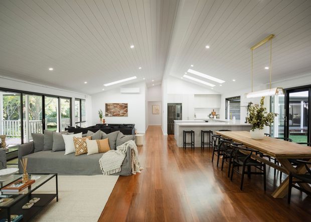 Bright, open-plan residential interior combining a living room, dining area and kitchen. The space features warm timber flooring, a large grey sectional sofa, a long timber dining table with black chairs, white cabinetry, extensive glazing, skylights and a white-lined ceiling. The room has a relaxed contemporary feel with abundant natural light.