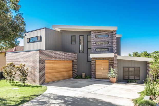 Exterior view of a contemporary two-storey home with dark brick masonry, vertical grey cladding, flat roof forms, narrow horizontal windows, timber garage door and timber entry door. A paved driveway, front lawn and established landscaping frame the facade.