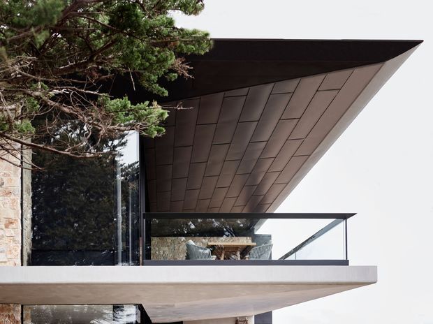 Close-up exterior architectural view of a contemporary building with a dramatic projecting cantilevered roof and balcony canopy. The soffit is clad in dark rectangular panels, with dark metal-framed glass balustrading and large reflective glazing. A natural stone wall is visible at the left, partially screened by mature trees.