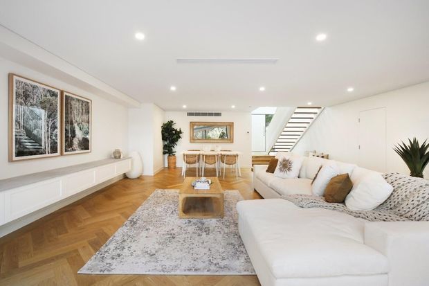 Bright open-plan living and dining area with a large cream sectional sofa, patterned area rug, white built-in media cabinetry, timber herringbone flooring, and a compact dining table. A contemporary open staircase, recessed ceiling lights, framed artwork, indoor plants, and neutral furnishings create a clean, warm, refined interior.