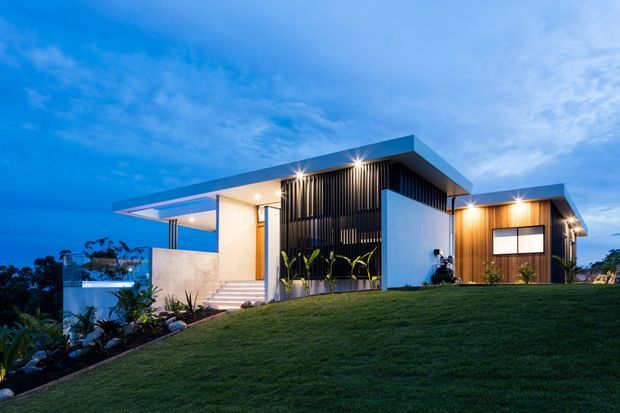 A contemporary single-storey residence photographed at dusk, set on a gently sloping landscaped site. The home features crisp white rendered volumes, a broad flat roof with deep cantilevered eaves, dark vertical screening, extensive glazing, warm timber cladding, and an illuminated entry terrace. A manicured lawn and planted garden frame the modern facade.