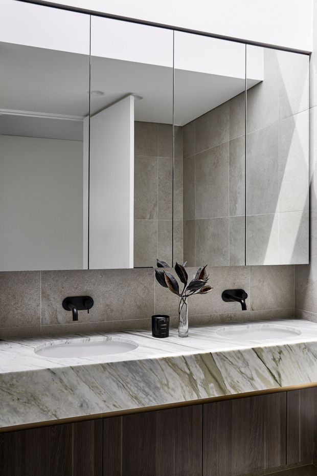 Contemporary bathroom vanity with a double integrated basin, dramatic veined stone benchtop, dark wood cabinetry, black wall-mounted taps, large mirrored storage, and grey stone-look wall tiles. Natural light creates a refined, high-contrast finish.