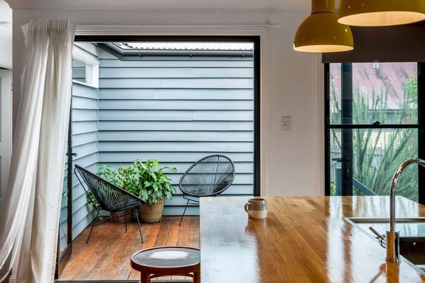 A bright contemporary kitchen opening onto a small enclosed outdoor courtyard or balcony. The kitchen features a large polished timber island with an undermount sink and curved chrome tap, black-framed glazing, and mustard pendant lights. Timber decking, pale blue-grey horizontal cladding, potted greenery, and black woven outdoor chairs are visible beyond the opening.