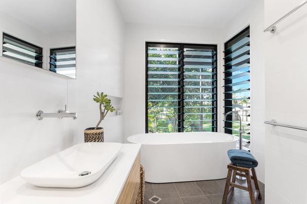 Bright contemporary bathroom featuring a freestanding white bathtub beneath black louvred windows, a wall-mounted basin on a white vanity, large mirror, dark grey tiled floor, and polished chrome fixtures. A small wooden stool and plant add warmth.