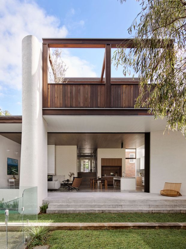 A contemporary two-storey residence viewed from the garden, featuring a covered open-plan living and kitchen area, broad stone steps, a tall white rendered cylindrical column, dark timber balcony cladding and balustrade, and large glazed openings. A glass pool or water-edge barrier is visible in the foreground, with established planting around the lawn.