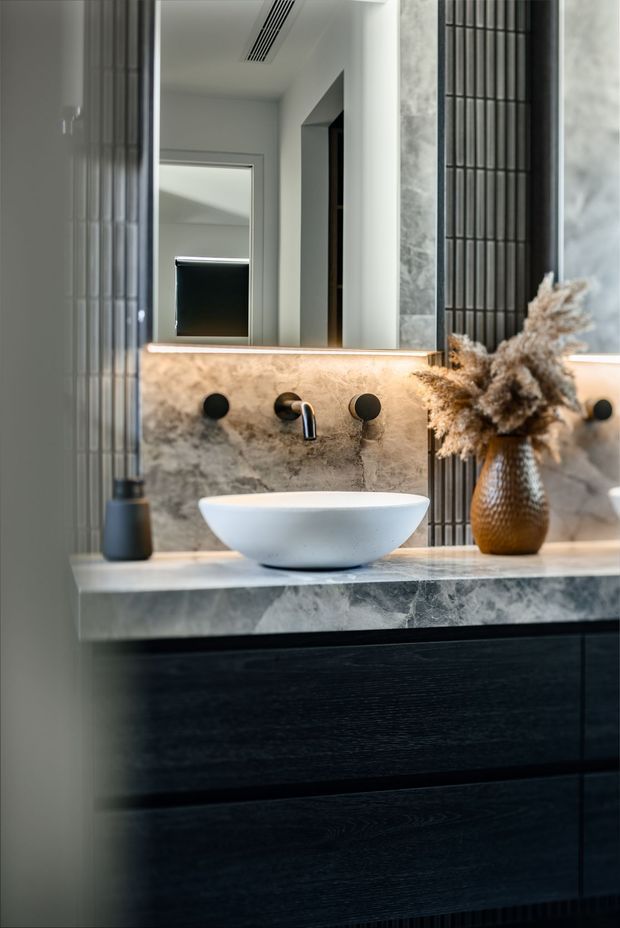 A close-up view of a refined contemporary bathroom vanity. A white vessel basin sits on a dark timber-look floating vanity with a grey veined stone benchtop and matching stone splashback. The wall-mounted black tapware and round handles contrast with vertical dark fluted wall panels, while a large mirror and warm integrated lighting add depth and atmosphere. A textured vase with dried foliage provides a decorative accent.
