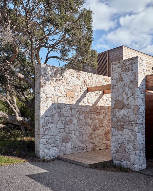 Exterior architectural view of a low, sculptural structure set among mature trees. Rough natural-stone walls form a courtyard-like entrance, complemented by warm horizontal timber cladding, timber beams, and a raised timber deck. The composition emphasizes the contrast between heavy masonry and lightweight timber, with a paved approach and landscaped surroundings under a partly cloudy sky.
