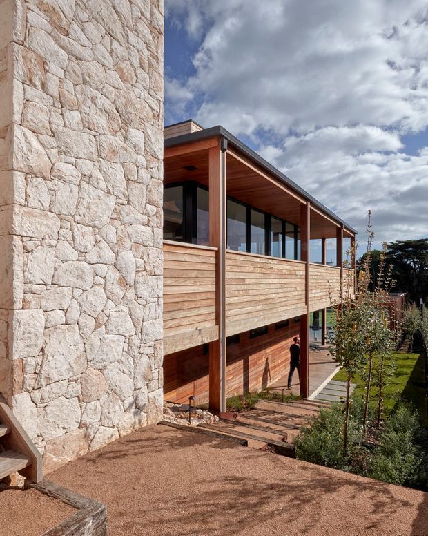 Exterior view of a contemporary multi-level residence built into a landscaped site. A prominent pale natural-stone wall contrasts with horizontal timber cladding, timber soffits, dark-framed glazing, and a projecting upper-level terrace or balcony. Steps and paths lead through a planted garden beside the house under a partly cloudy sky.