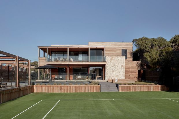 Two-storey contemporary residence with a warm timber-clad facade, expansive glazed balconies and ground-floor openings, and a prominent natural stone feature wall. The house fronts a landscaped artificial grass tennis court, with timber decking, fencing, and outdoor terraces visible around the building.