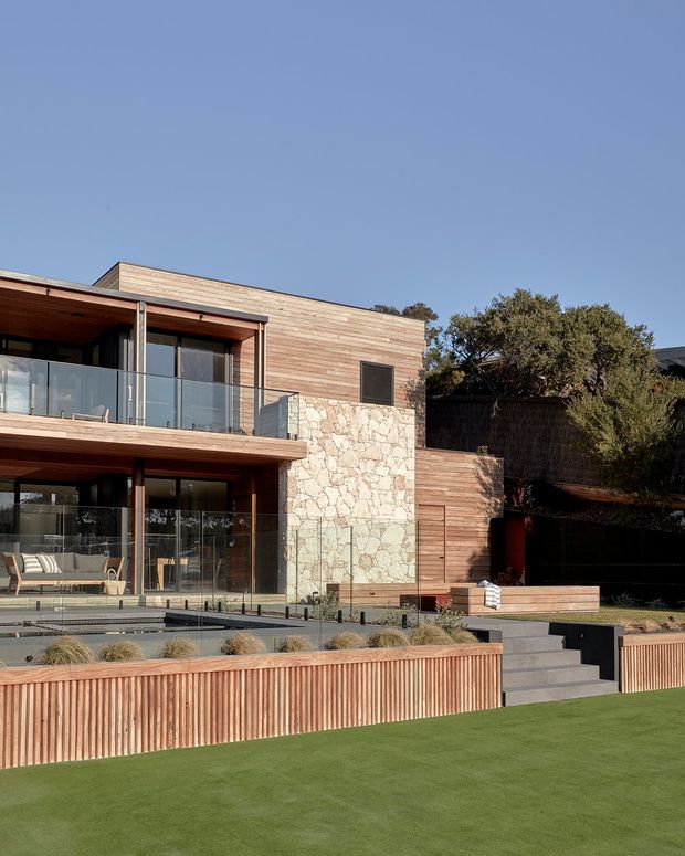 A contemporary multi-level residential facade with extensive horizontal timber cladding, large floor-to-ceiling glazed doors and windows, and a prominent natural stone feature wall. The building has recessed balconies with clear glass balustrades, timber soffits, stepped terraces, landscaped planting, and a neatly finished lawn in the foreground. Warm timber and stone contrast with dark glazing and the surrounding mature vegetation.