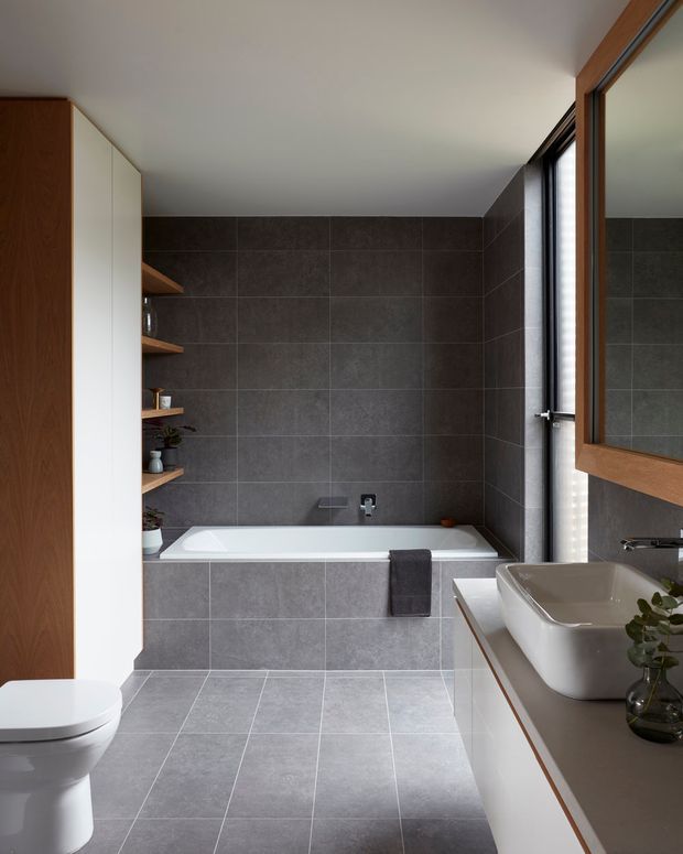 A modern bathroom with a grey tiled floor and walls, a built-in white bathtub, wall-mounted fixtures, a white toilet, and a floating vanity with a white vessel basin. Warm timber cabinetry and a large timber-framed mirror add contrast, while open timber shelving and a narrow window bring detail and natural light into the room.