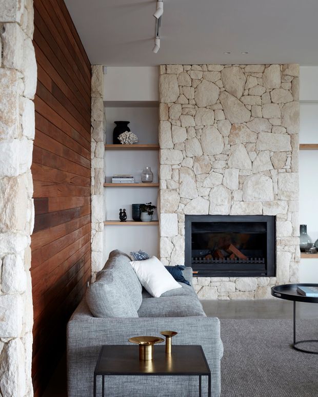 A contemporary living room centred on a substantial pale natural-stone fireplace, with built-in open shelving, a grey sectional sofa, dark coffee tables, and warm horizontal timber wall panelling. The room has a restrained neutral palette, clean-lined furniture, and recessed or track-style ceiling lighting.