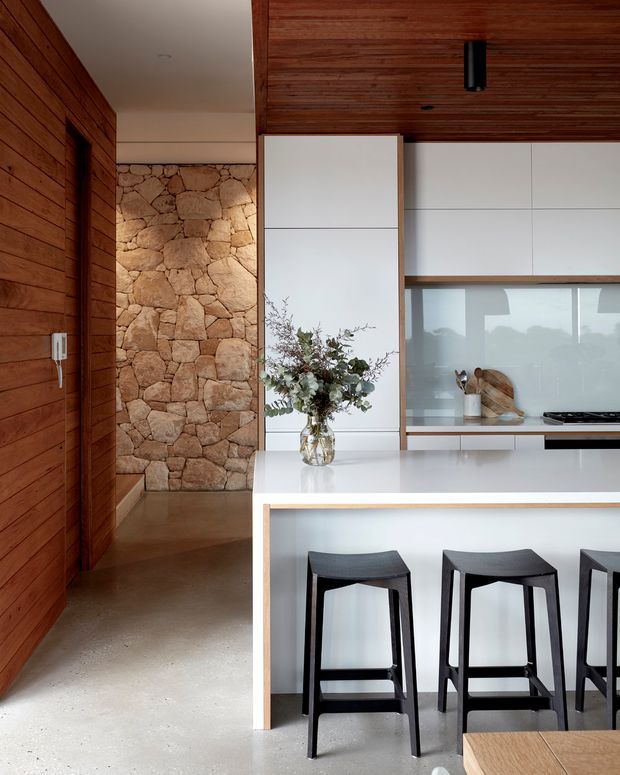 A contemporary kitchen with a white island and integrated breakfast bar, three dark stools, white flat-panel cabinetry, and a concealed or glazed backsplash area. Warm timber wall and ceiling lining contrast with a rough natural stone feature wall and polished concrete floor. The space has clean lines, understated black lighting, and abundant natural light.