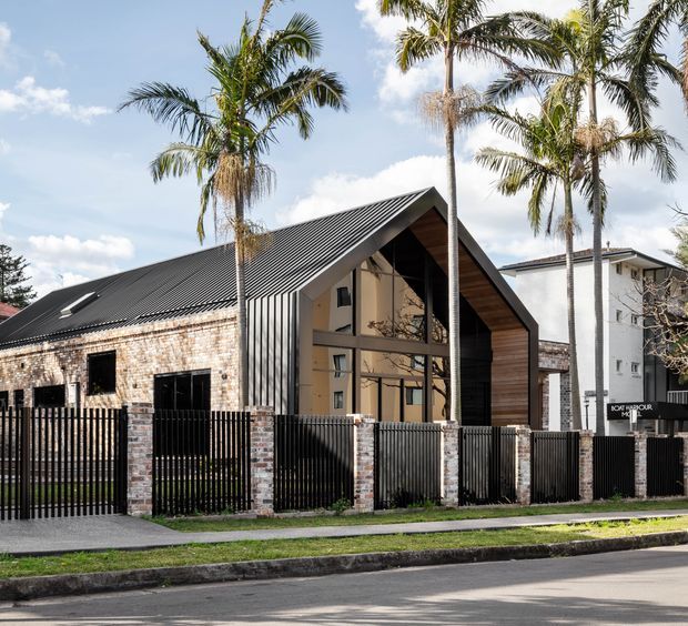 Street-facing photograph of a contemporary gabled building with a steep dark metal roof, exposed brick walls, timber-clad gable frontage, and large glazed openings. A black vertical-slat fence with brick piers defines the front boundary, while tall palms and neighbouring buildings surround the property.