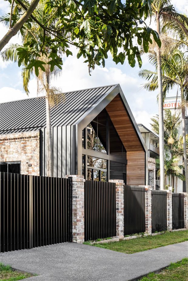 Street-facing view of a contemporary residential building with a steep gabled roof, dark standing-seam metal roofing and cladding, warm timber-lined gable, extensive glazing, and exposed brick walls. A tall black slatted fence with brick piers fronts the property, with palms and tropical vegetation around it.