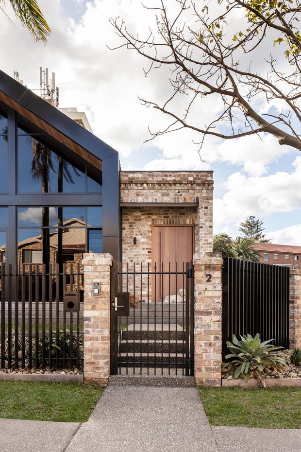 Street-facing view of a contemporary residential facade combining dark metal framing, expansive reflective glazing, warm timber detailing, and a weathered brick entry pavilion. A brick-and-black-metal fence with pedestrian gate leads to the timber front door, with a concrete path and sparse landscaped planting in the foreground.