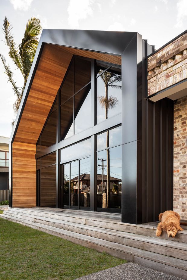 Exterior view of a contemporary residential extension or facade with a dramatic angular black metal frame, warm timber-lined soffit and wall, large floor-to-ceiling glazing, and a contrasting exposed brick volume. Broad timber steps lead to the glazed entrance, with a small lawn and paved foreground.