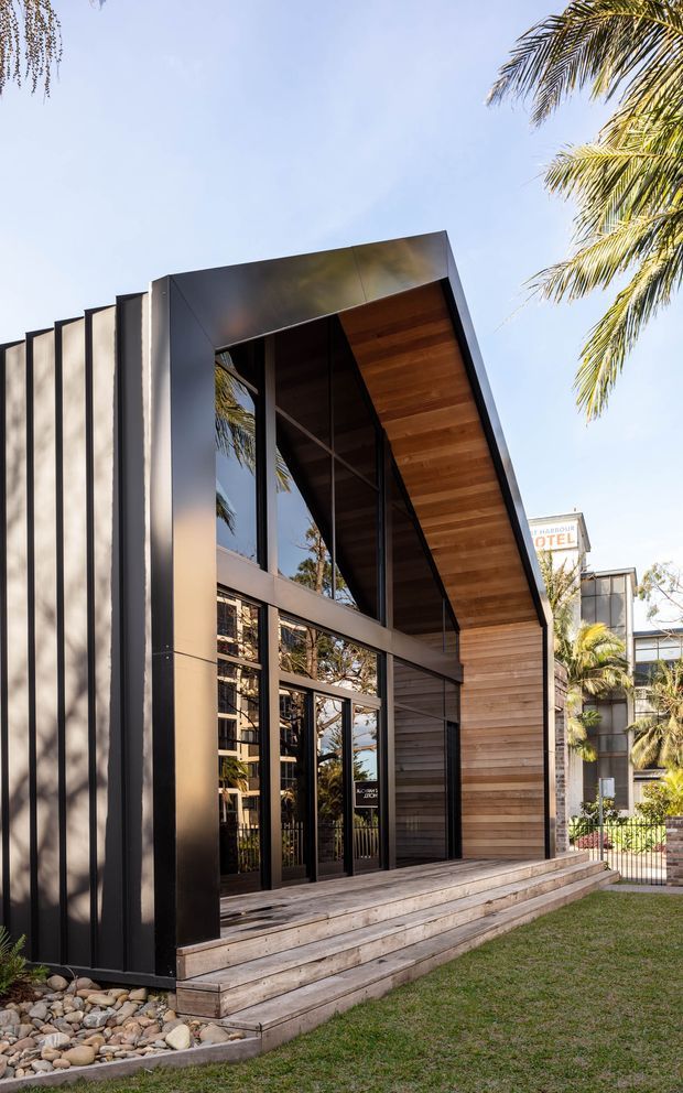 Exterior view of a contemporary hotel entrance with a tall, angular portal clad in dark metal and warm horizontal timber. Floor-to-ceiling glazed entry doors and windows sit beneath a timber-lined projecting canopy, with stepped timber decking, landscaping, and palm trees around the entrance.