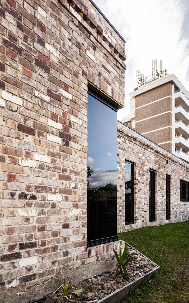 Exterior architectural view of a low-rise brick building with varied reclaimed-looking brickwork, tall dark-framed reflective glazing, and a simple landscaped strip of river stones and grass. A larger brick apartment building is visible in the background.