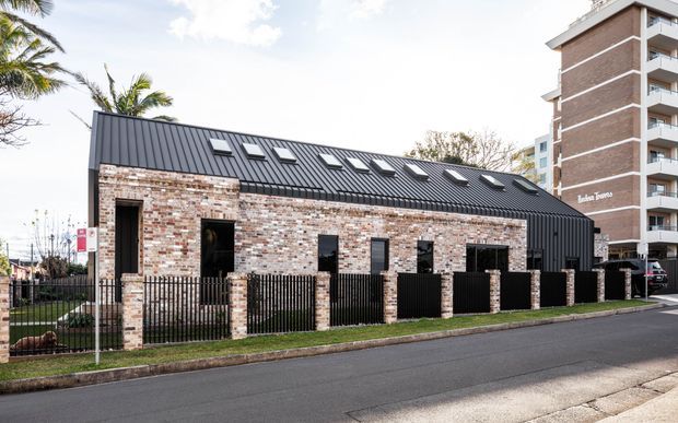 Exterior view of a contemporary low-rise brick building with a long pitched dark metal roof, multiple roof skylights, black-framed openings, and a black metal front fence. A taller apartment building is visible behind it, with palm trees and a suburban streetscape surrounding the site.