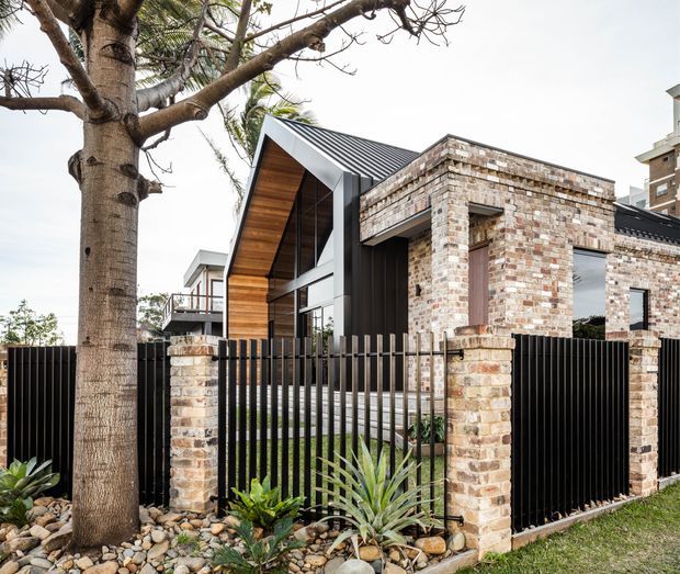 Contemporary residential house exterior viewed from the street, combining exposed reclaimed-looking brickwork, black vertical metal fencing, dark standing-seam roofing, timber-lined gable soffits, and extensive glazing. A landscaped verge with a mature tree, river stones, and drought-tolerant planting fronts the property.