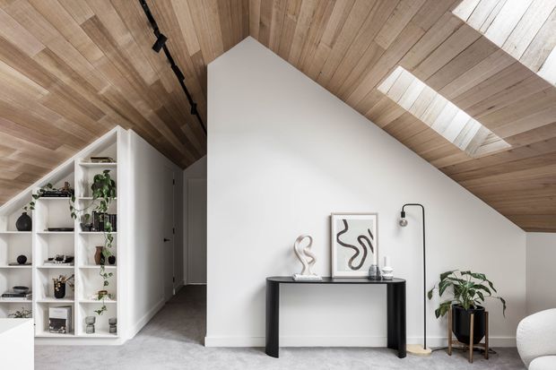 Contemporary attic-style living space with a dramatic sloped timber-lined ceiling, skylights, white walls, and a minimalist black console styled with artwork and plants. Built-in open shelving and a hallway are visible to the left, with a grey carpeted floor and restrained neutral furnishings.