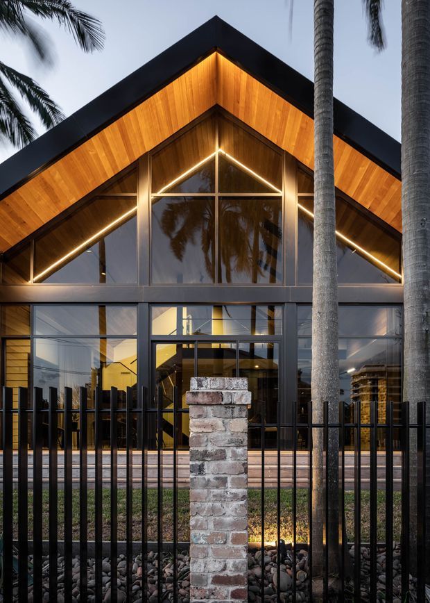 Front exterior view of a contemporary gabled building at dusk, featuring a dramatic timber-lined triangular roof facade, dark metal framing, extensive glazing, integrated linear lighting, and a brick entry pier behind a black metal fence. Palm trees frame the facade, and warm interior lighting creates a welcoming appearance.