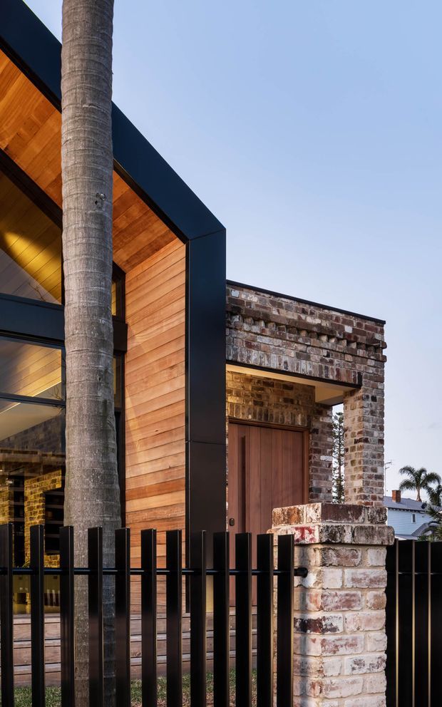 A close-up exterior architectural view of a contemporary home featuring warm horizontal timber cladding and soffits, dark metal framing, extensive glazing, and a contrasting weathered brick entry surround with a timber door. A palm trunk and black vertical metal boundary fencing frame the foreground.
