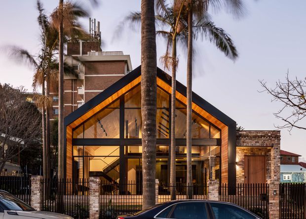 Exterior street view of a contemporary, steeply gabled building with extensive floor-to-ceiling glazing, dark structural framing, and warm timber lining beneath the roof. Tall palm trees partially screen the facade, while a taller brick building and neighbouring structures sit behind it. The illuminated interior is visible through the glass, including a stairway and open-plan spaces.