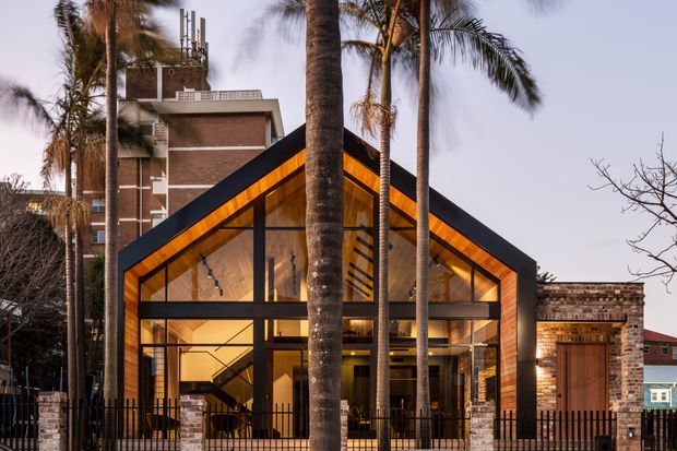 Front exterior view of a contemporary gable-front house framed by tall palm trees. The home features a steeply pitched roof with dark metal framing, extensive floor-to-ceiling glazing, warm timber-lined soffits, exposed brick walls, and a black metal fence. A taller brick building is visible behind it.