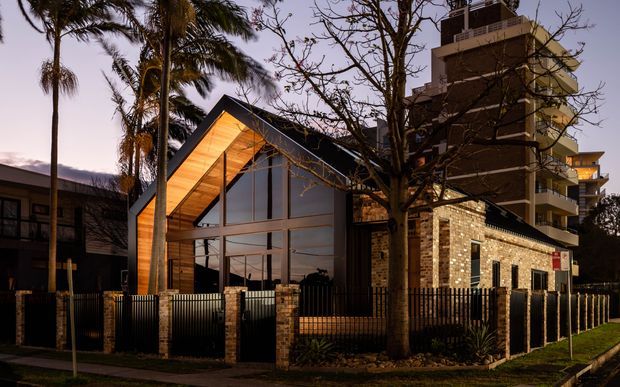 Twilight exterior view of a contemporary multi-storey residential development combining a prominent timber-lined gable volume with extensive glazing and a taller brick apartment building behind it. Palm trees, a mature street tree, black metal fencing, balconies, and warm interior lighting frame the urban streetscape.