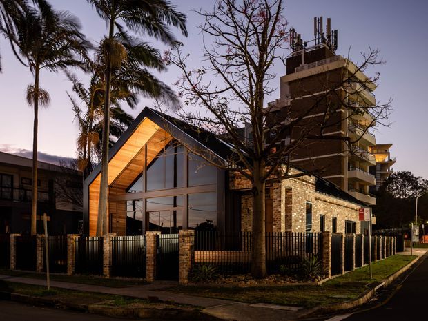 Twilight street view of a contemporary building with a dramatic steeply pitched, timber-lined gable roof and extensive floor-to-ceiling glazing. The facade combines warm timber, dark metal framing, and exposed light brick, with a taller apartment building behind it. A fenced landscaped verge, mature palm trees, and street frontage are visible.