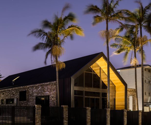 Twilight exterior view of a contemporary gable-roof building with a strongly expressed timber-lined triangular entry facade, dark metal roofing, extensive glazing, brick masonry walls, and a black vertical-slat fence. Tall palm trees frame the building, with a neighboring multi-storey structure visible behind it.