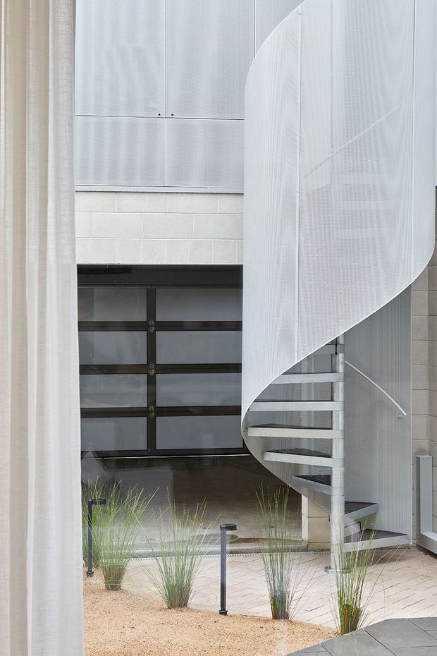 A bright contemporary courtyard or entry space featuring a sculptural white perforated-metal spiral staircase, a dark-framed sectional garage door, pale concrete block walls, and light paving bordered by ornamental grasses and gravel. A sheer curtain partially frames the view.