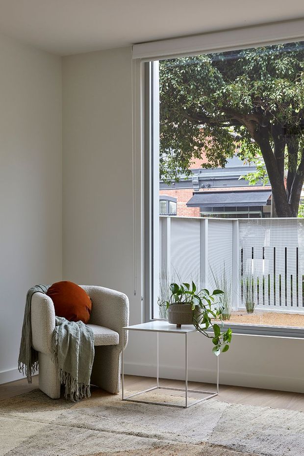 A bright, calm sitting area with a textured cream boucle armchair, rust-orange cushion, draped sage-grey throw, small white side table, potted trailing plant, and a large floor-to-ceiling window overlooking a planted courtyard and neighbouring brick building. Neutral walls, light timber flooring and a pale textured rug create a soft, minimalist interior.