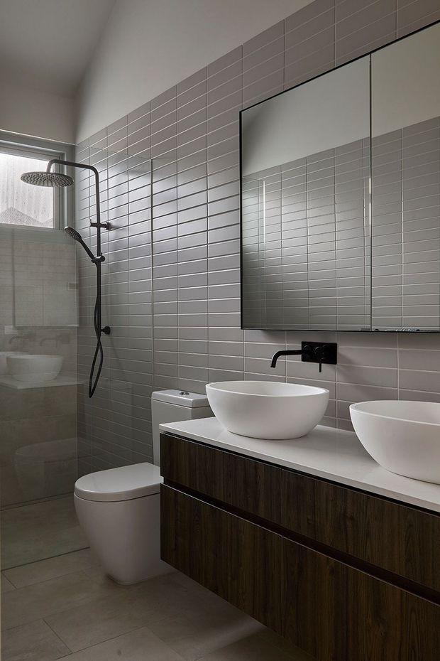 A contemporary bathroom with a walk-in glass shower, wall-mounted rain shower and handheld fixture, wall-hung toilet, double vessel basins, large mirrored cabinet, dark timber-look vanity cabinetry, and a white benchtop. The room is finished with pale grey horizontal rectangular wall tiles and matching large-format floor tiles.