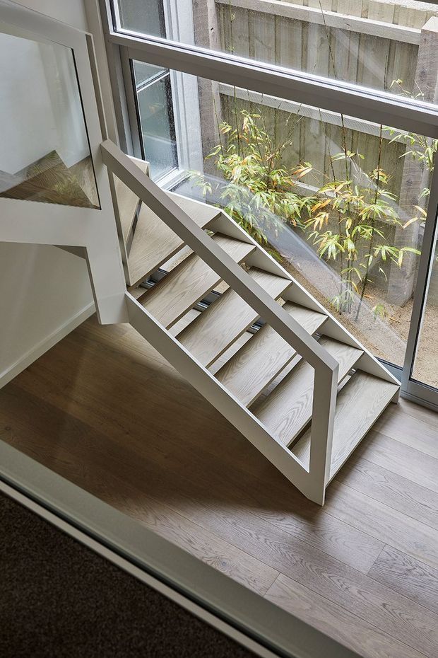 Interior view of a compact, contemporary stairway with pale timber open-riser steps and a simple painted white railing. The staircase sits beside large floor-to-ceiling windows, overlooking a narrow planted courtyard. Light timber flooring, white walls, and abundant daylight create a clean, airy atmosphere.