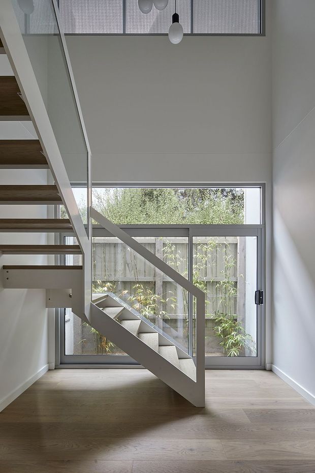 A bright, minimalist stairway with open-riser timber treads and a white-painted structural balustrade. The stairwell has a high ceiling, pale timber flooring, white walls, large aluminium-framed glazing, and a glazed rooflight or clerestory above. A planted courtyard is visible through the windows, and simple pendant lights hang overhead.
