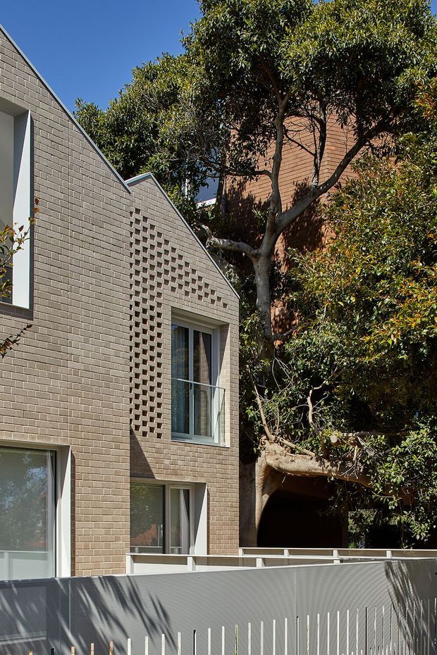 Exterior view of a contemporary multi-storey residence with a steeply pitched gable form, pale grey-brown brick walls, recessed windows, and a patterned breeze-block brick screen. A screened terrace or boundary fence occupies the foreground, with mature trees closely surrounding the building.