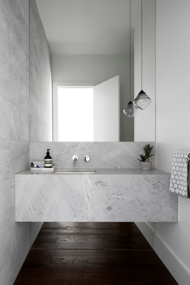 A compact contemporary bathroom featuring a floating stone vanity with integrated basin, large wall mirror, pale marble-look wall and vanity surfaces, dark timber flooring, minimalist chrome tapware, pendant lights, and a small potted plant. A doorway and towel rail are visible in the background and at right.