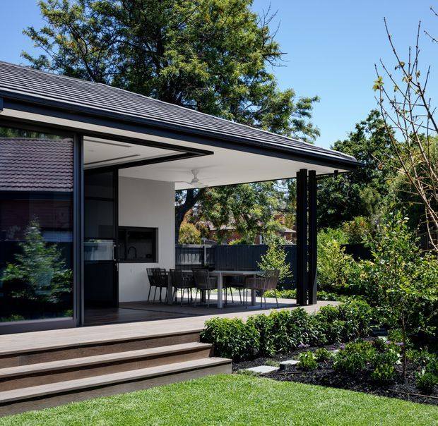 A contemporary single-storey residential extension or pavilion viewed from the garden, with a broad covered outdoor dining deck, slim dark structural posts, expansive dark-framed glazing, white walls, and a low-pitched corrugated metal roof. Timber steps lead from the deck to a manicured lawn and planted garden. A compact kitchen and dining area are visible beneath the covered roof.
