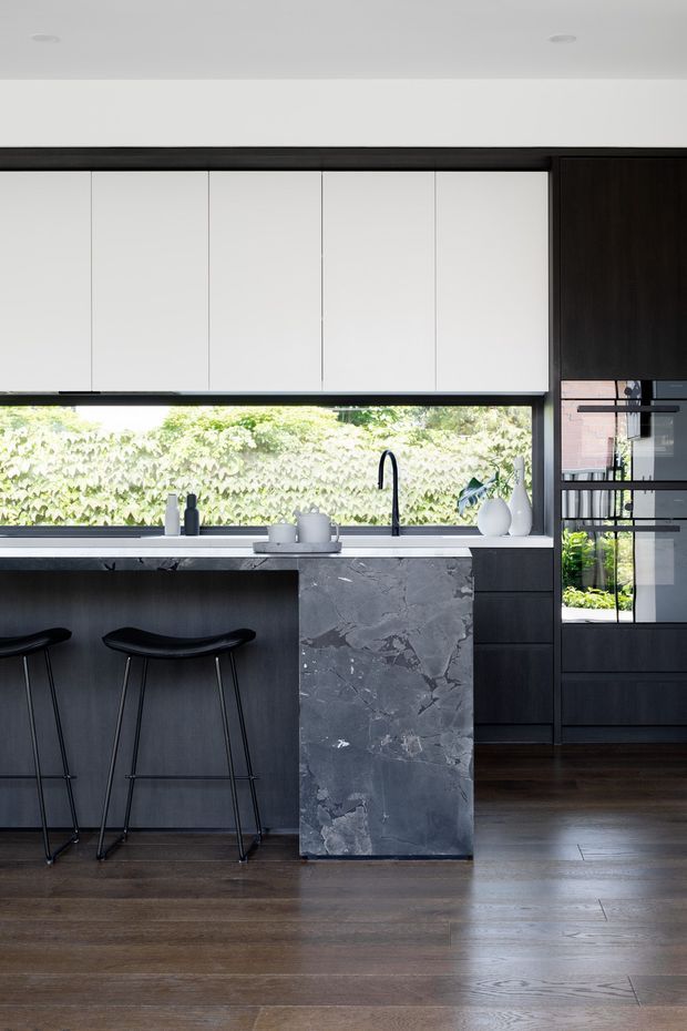 A contemporary kitchen with a dark timber island and cabinetry, white overhead cupboards, a dramatic grey stone island end panel, and a long horizontal window overlooking greenery. Black bar stools sit at the island, while integrated dark-framed glazing and appliances are visible to the right. The room has dark timber flooring and a refined, high-contrast palette.