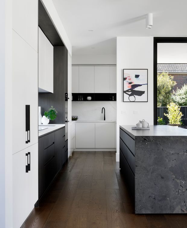 A contemporary galley-style kitchen with a large central island. The space features flat-panel white cabinetry, dark timber or charcoal cabinetry, black linear handles, a dark stone island benchtop and waterfall end, a white tiled or stone splashback, timber flooring, and black-framed glazing opening to an exterior balcony or garden. A small pantry or secondary kitchen zone is visible at the rear.